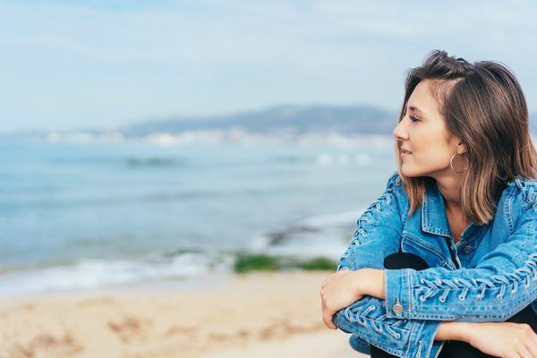 Thoughtful young woman gazing out over the sea as she sits in a denim jacket above a sandy beach on a misty day